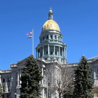 An image displaying the Colorado State Capitol building, featuring the state flag flying atop against a backdrop of a clear blue sky.