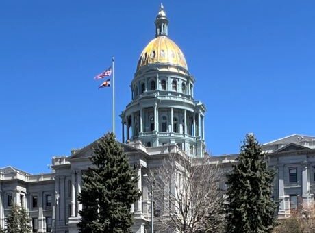 An image displaying the Colorado State Capitol building, featuring the state flag flying atop against a backdrop of a clear blue sky.