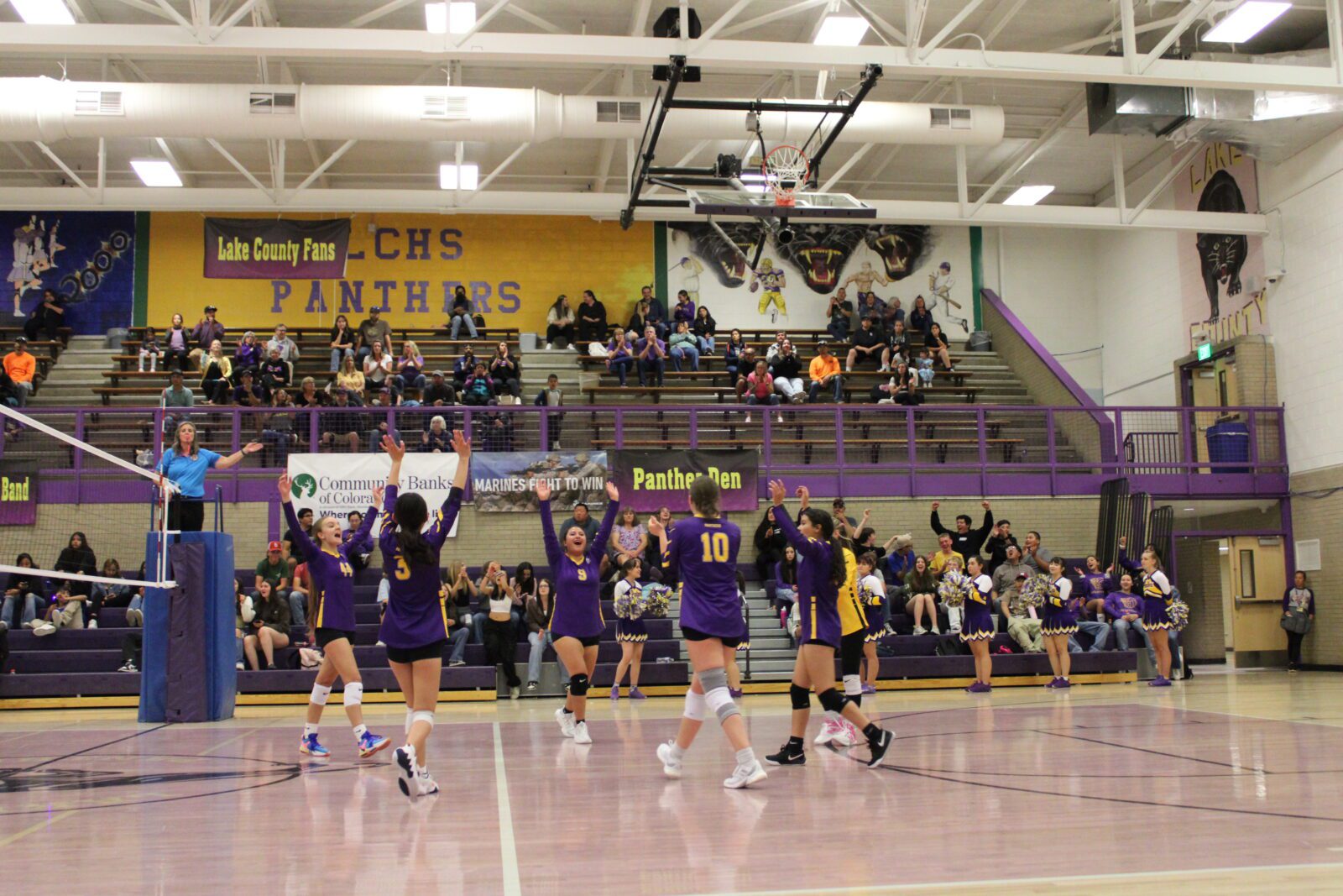 Group of students playing volleyball inside a school gymnasium, with several players in action and student watching in the back.